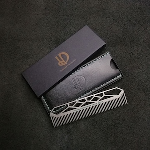 May include: A silver comb with a geometric design and the text "Наположенное сила духа" in black lettering. The comb is in a black leather case with a stitched border.