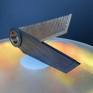 May include: A black wooden comb with a silver metal comb attached to it. The comb is open and the metal comb is facing up. The comb is sitting on a white surface.