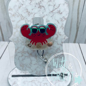May include: A red crab with green sunglasses and a white background. The crab is wearing sunglasses and is surrounded by seashells. The crab is on a white marble background with a black base.