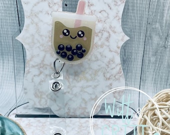 May include: A clear plastic badge holder with a brown and white cartoon boba tea charm. The charm has a smiling face and black boba pearls. The holder has a silver clip and two silver snaps. The holder is on a marble background.