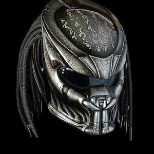 Può includere: Un casco da moto argentato con un design Predator. Il casco presenta una superficie dettagliata e strutturata, una visiera scura e fili simili a dreadlock. Il design include motivi intricati e un aspetto minaccioso e alieno.