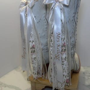 Puede incluir: Botas de vaquero grises claras con detalles florales y perlas. Cada bota está adornada con un lazo y una cinta blancos, con el nombre "Mrs. Kellogg" impreso en la cinta. Las botas se exhiben sobre un pequeño soporte de madera.