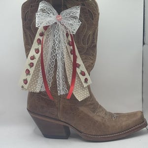 Może przedstawiać: Brązowy sk&oacute;rzany but kowbojski z ozdobną kokardą. Kokarda posiada białą koronkę, czerwone wstążki oraz wstążki z wzorem truskawek i groch&oacute;w. But ma kwadratowy nosek i warstwowy obcas.