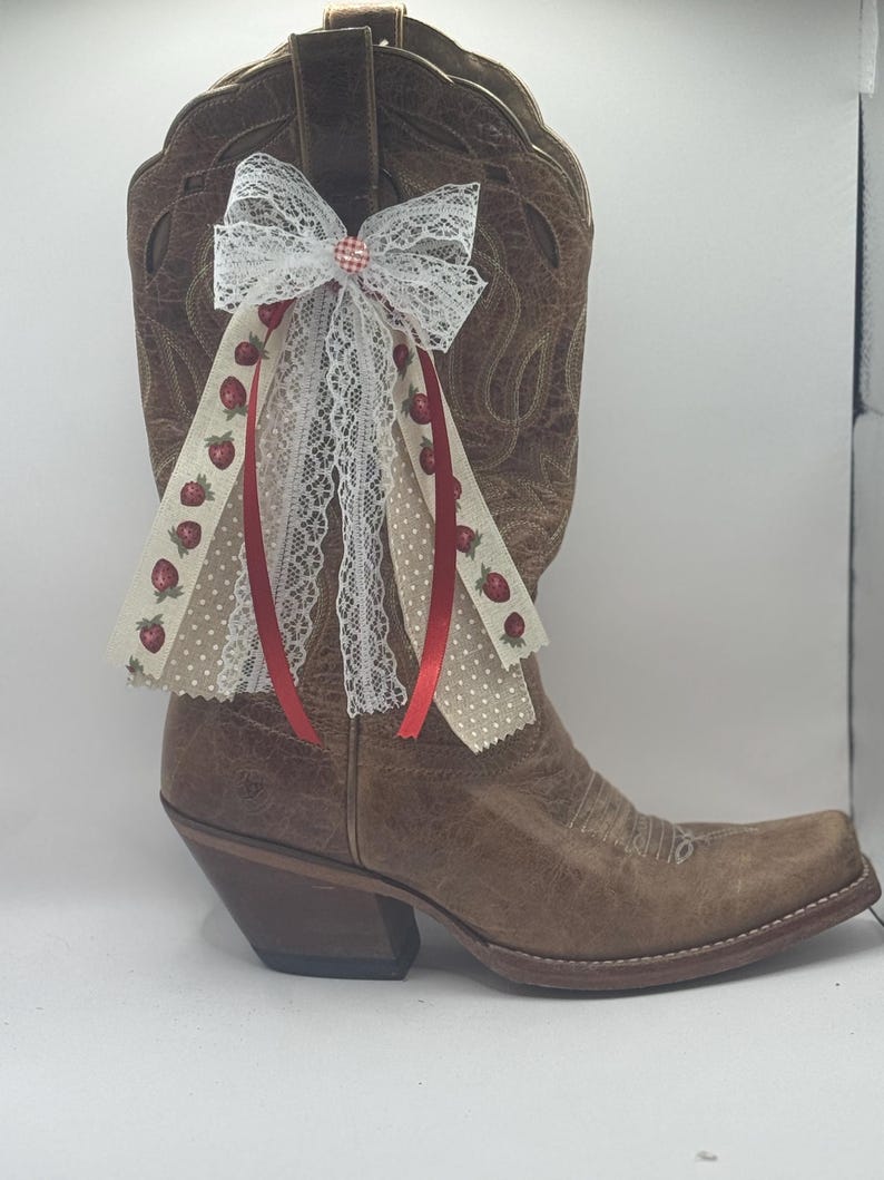 Może przedstawiać: Brązowy sk&oacute;rzany but kowbojski z ozdobną kokardą. Kokarda posiada białą koronkę, czerwone wstążki oraz wstążki z wzorem truskawek i groch&oacute;w. But ma kwadratowy nosek i warstwowy obcas.