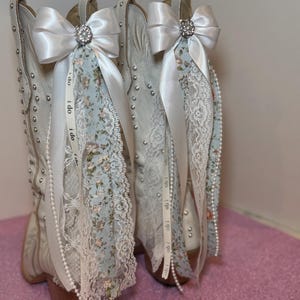 Pode incluir: Um par de botas de cowboy brancas adornadas com laços de cetim branco, enfeites de strass e detalhes de renda floral. As botas apresentam tachas prateadas e fitas com a inscrição "I do".
