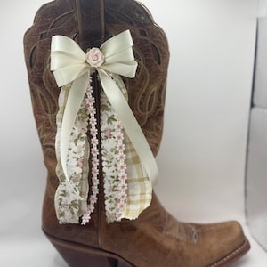 May include: A brown leather cowboy boot adorned with a cream-colored bow, a small pink rose, and floral and plaid ribbons. The boot has a pointed toe and a stacked heel, showcasing a rustic, country aesthetic.