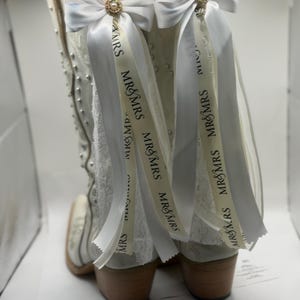 Op de afbeelding: Witte cowboylaarzen met decoratieve versieringen. Elke laars heeft een wit lint met de tekst "MR&MRS" en een decoratieve strik met een goudkleurig juweel.
