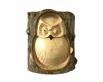May include: A carved wooden owl sculpture, painted in a light brown color, is set into a piece of tree bark. The owl has large, round eyes and a detailed carving of its feathers.