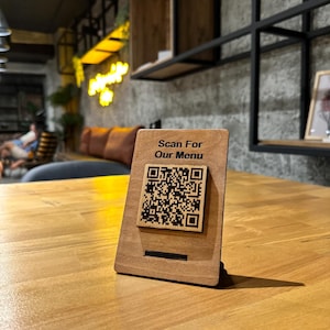 Peut inclure: Un présentoir de table en bois avec le texte "Scan For Our Menu" et un code QR. Le présentoir est en bois brun clair et est posé sur une table en bois. L'arrière-plan montre un cadre de restaurant.