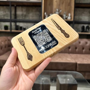 Peut inclure: Un porte-menu rectangulaire en bois avec des motifs gravés de fourchette et de couteau. Un autocollant noir avec un code QR et le texte "Scan For Menu" est apposé. Le support est marqué "DECORIUM WOOD."