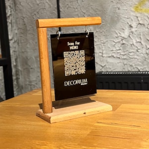 Peut inclure: Un présentoir de table en bois avec un panneau en acrylique noir. Le panneau affiche un code QR et le texte "Scan For MENU" et "DECORIUM WOOD-". Le présentoir est en bois clair et est conçu pour être posé sur une table.