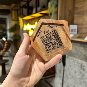 May include: A wooden hexagonal menu holder with a QR code and the words "Scan For Menu" engraved on its surface. The holder is made of light-colored wood and is held in a person's hand.
