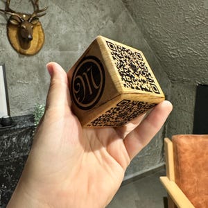 Può includere: Un cubo di legno con codici QR su più lati. Un lato presenta il numero "916" in un design circolare. Il cubo è tenuto in mano, mostrando la venatura naturale del legno e gli intricati dettagli dei codici QR.