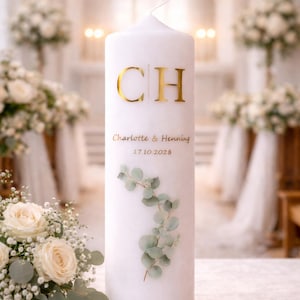 Könnte beinhalten: Eine weiße Säulenkerze mit goldenen Buchstaben und einem Zweig grüner Blätter. Die Kerze trägt die Initialen "CH", die Namen "Charlotte & Henning" und das Datum "17.10.2028". Die Kerze ist wahrscheinlich für eine Hochzeit oder ein besonderes Ereignis.