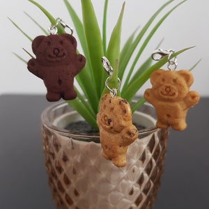 May include: Three small bear-shaped charms, one brown, one light brown, and one tan, hanging from silver chains. The charms are hanging from a green plant in a clear glass vase.