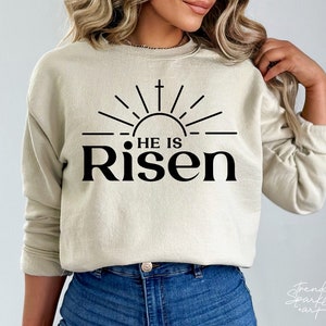May include: A beige sweatshirt with a black graphic design of a sun with a cross and the text "He is Risen".