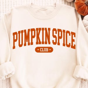 May include: A cream-coloured crewneck sweatshirt featuring the text PUMPKIN SPICE CLUB in a bold, arched orange font. The word PUMPKIN SPICE is printed in large capital letters, while the word CLUB is inside an orange oval with two dots.