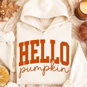 May include: A cream-colored hooded sweatshirt featuring the text HELLO pumpkin printed in a bold, burnt orange font. The word HELLO is in a large, block-style typeface, while the word pumpkin is in a smaller, cursive-style script font.