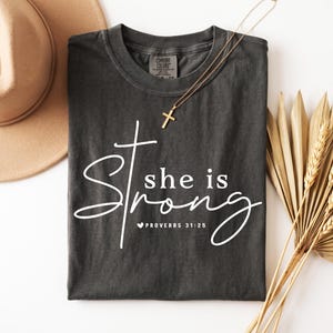 May include: Dark gray t-shirt with the words "she is Strong" in white script, a cross graphic, and the Bible verse Proverbs 31:25. A gold cross necklace and a tan hat are also in the image.