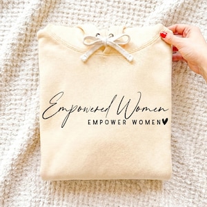 May include: A cream-coloured hoodie featuring the words "Empowered Women Empower Women" in black script. The hoodie has a drawstring and is neatly folded. A hand with red nail varnish holds the garment.