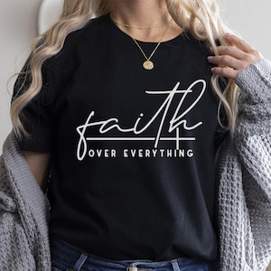 May include: A black t-shirt with white text that reads "faith over everything". The shirt is being worn by a person with long blonde hair and a gray cardigan.