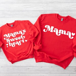 May include: Two red sweatshirts with white text. The larger sweatshirt says "Mama" in a retro font with a heart. The smaller sweatshirt says "Mama's Sweetheart" in a retro font with a heart.
