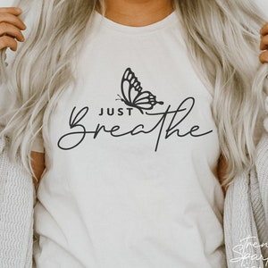 Puede incluir: Camiseta color crema con las palabras "Just Breathe" en una fuente elegante. Un gráfico de mariposa negra está encima de la palabra "Breathe". La camiseta es de color neutro y tiene un ajuste relajado.