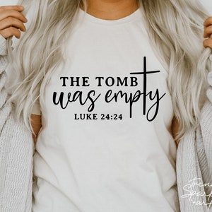 May include: Cream-colored t-shirt with the text "THE TOMB was empty" and a cross graphic. The Bible verse Luke 24:24 is printed below the text in a black, elegant font. The shirt is a crew neck style.