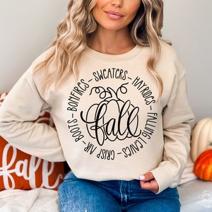 May include: A beige sweatshirt with a black graphic of a pumpkin and the word "fall" surrounded by text that reads "Crisp Air - Boots - Bonfires - Sweaters - Hayrides - Falling Leaves".