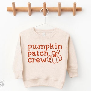 Puede incluir: Una sudadera de manga larga beige con el texto "Pumpkin Patch Crew" en naranja y un pequeño gráfico de calabaza naranja.