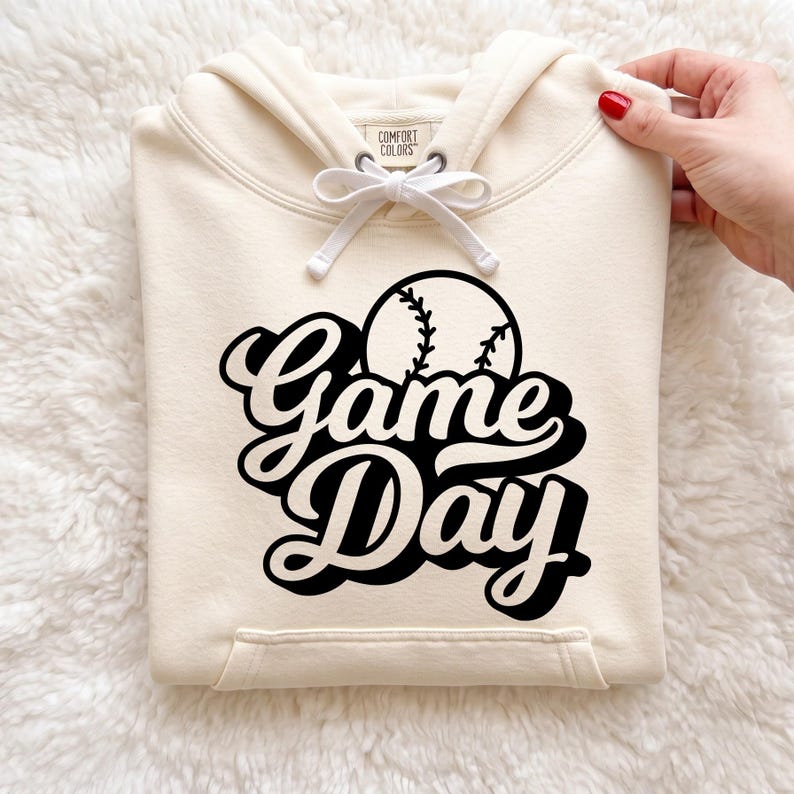 May include: A cream-colored hooded sweatshirt featuring a black graphic print of a baseball and the text "Game Day" in a bold, cursive font. The hoodie includes a matching cream drawstring and a Comfort Colors brand label at the neckline.