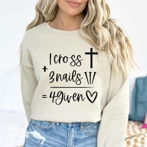 May include: A beige crew neck sweatshirt featuring the text "1 cross + 3 nails W/ = 4given" in black script, with a cross and heart symbol. The sweatshirt is a neutral colour.