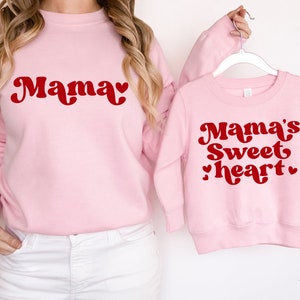 May include: Two pink sweatshirts with red text. The larger sweatshirt says "Mama" with a heart. The smaller sweatshirt says "Mama's Sweetheart" with two hearts.