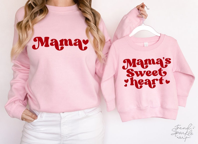 May include: Two pink sweatshirts with red text. The larger sweatshirt says "Mama" with a heart. The smaller sweatshirt says "Mama's Sweetheart" with two hearts.