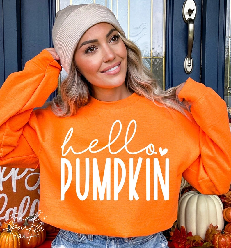 May include: An orange sweatshirt with the words "hello PUMPKIN" in white script. The sweatshirt is worn with a beige beanie. The image has a fall theme, with pumpkins and autumn decor in the background.