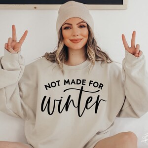 May include: A beige sweatshirt with the text "Not Made For Winter" in black lettering. The model is wearing a beige beanie and has their hands in the air.
