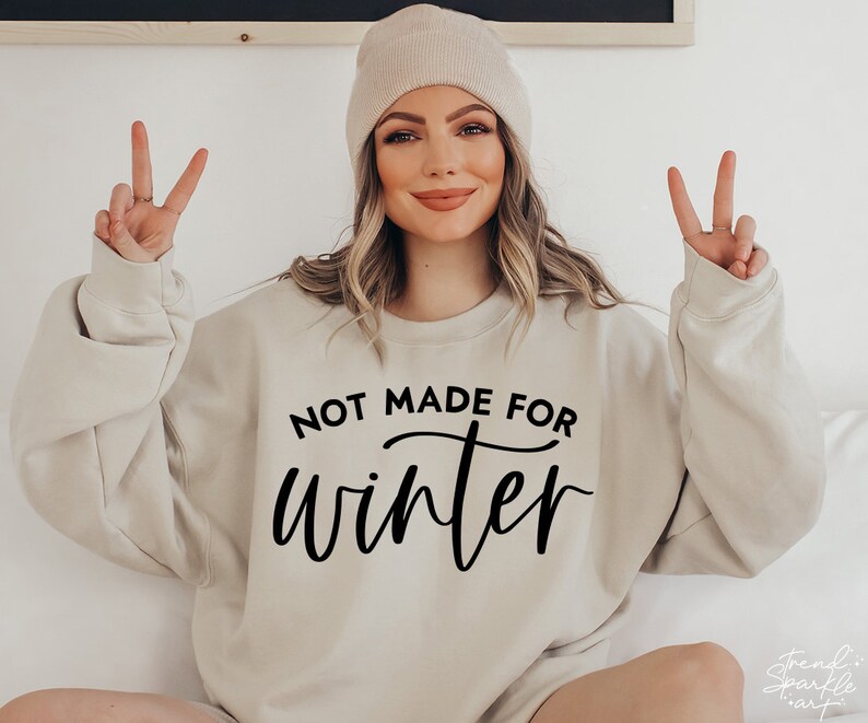 May include: A beige sweatshirt with the text "Not Made For Winter" in black lettering. The model is wearing a beige beanie and has their hands in the air.