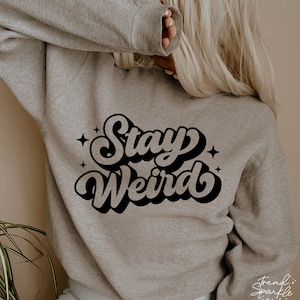 以下が含まれることがあります： グレーのトレーナーに、黒いレトロな書体で「Stay Weird」という文字が書かれています。文字は白で縁取られており、3D効果があります。