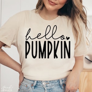 May include: A cream-coloured t-shirt with the words "hello PUMPKIN" in black script and block letters. A small heart is above the word "PUMPKIN". The shirt is a casual style.