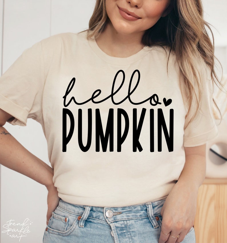 May include: A cream-coloured t-shirt with the words "hello PUMPKIN" in black script and block letters. A small heart is above the word "PUMPKIN". The shirt is a casual style.