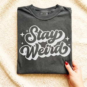 May include: Dark gray t-shirt with the words "Stay Weird" in a white, retro-style font. The text is surrounded by small starbursts. The shirt is made of a soft material and has a crew neck.