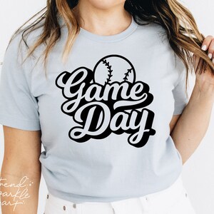 May include: A light blue short-sleeved t-shirt featuring a black graphic print on the chest. The design includes a baseball illustration positioned above the bold, stylized black text that reads Game Day in a cursive, layered font style.