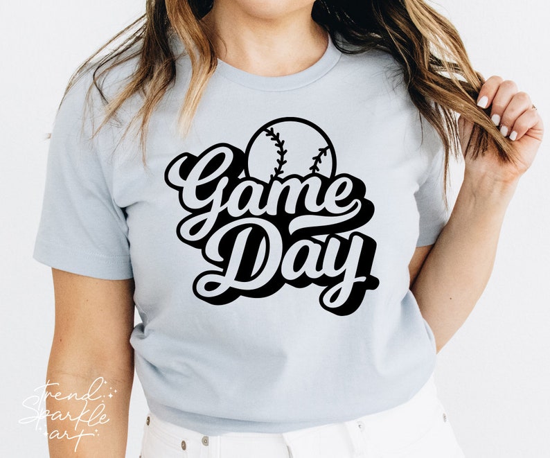 May include: A light blue short-sleeved t-shirt featuring a black graphic print on the chest. The design includes a baseball illustration positioned above the bold, stylized black text that reads Game Day in a cursive, layered font style.