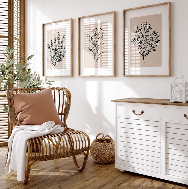 Puede incluir: Un armario blanco con dos puertas y dos cajones, una silla de mimbre con un coj&iacute;n marr&oacute;n y tres impresiones enmarcadas de ilustraciones florales en blanco y negro en una pared blanca.