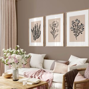 Puede incluir: Tres dibujos en blanco y negro de flores en marcos sobre una pared beige. Los marcos son de madera clara y la pared es de color marr&oacute;n claro. Un sof&aacute; blanco con almohadas y una manta de color rosa est&aacute; delante de la pared.