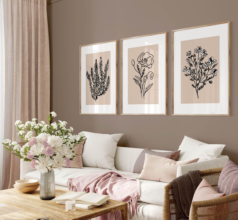 Puede incluir: Tres dibujos en blanco y negro de flores en marcos sobre una pared beige. Los marcos son de madera clara y la pared es de color marr&oacute;n claro. Un sof&aacute; blanco con almohadas y una manta de color rosa est&aacute; delante de la pared.