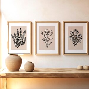 Puede incluir: Tres impresiones enmarcadas con dibujos de l&iacute;nea negra de diferentes flores sobre un fondo beige claro. Los marcos son de madera clara y est&aacute;n colgados en una pared blanca.