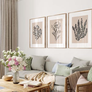 Puede incluir: Tres dibujos en blanco y negro de diferentes flores sobre un fondo beige, enmarcados. Los marcos son de madera clara y est&aacute;n colgados en una pared blanca sobre un sof&aacute; gris.