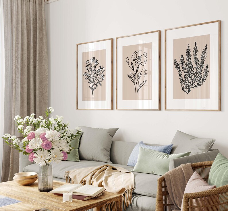 Puede incluir: Tres dibujos en blanco y negro de diferentes flores sobre un fondo beige, enmarcados. Los marcos son de madera clara y est&aacute;n colgados en una pared blanca sobre un sof&aacute; gris.