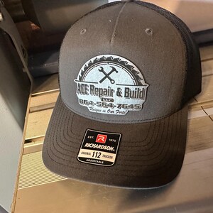May include: A black and gray trucker hat with a white logo that says "ACE Repair & Build LLC" with a saw blade and crossed wrenches. The hat is adjustable and has a red tag that says "Richardson" and "Original 112 Trucker".